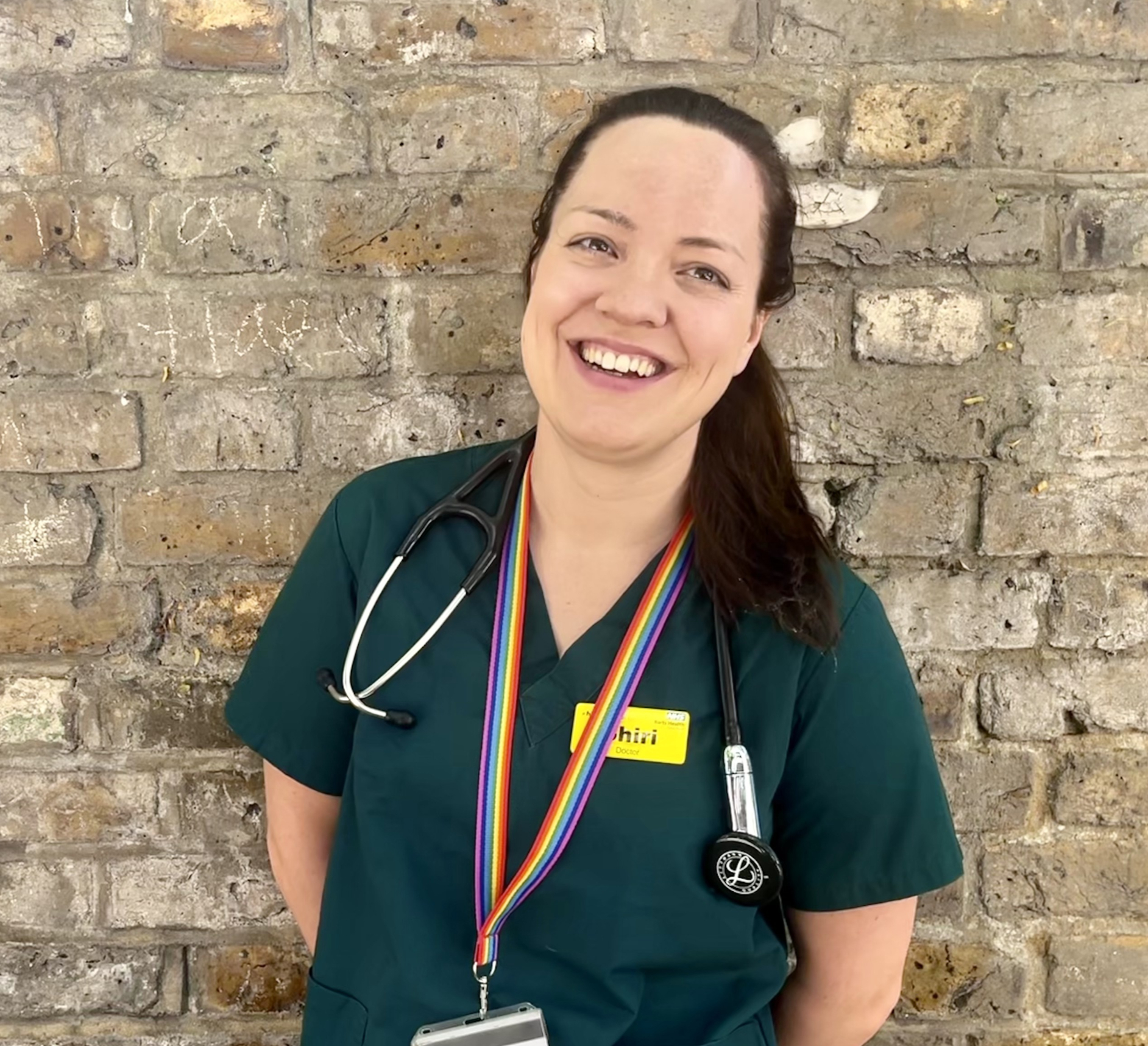 Dr Shiri Lejay standing in front of a brick wall wearing her scrubs and smiling